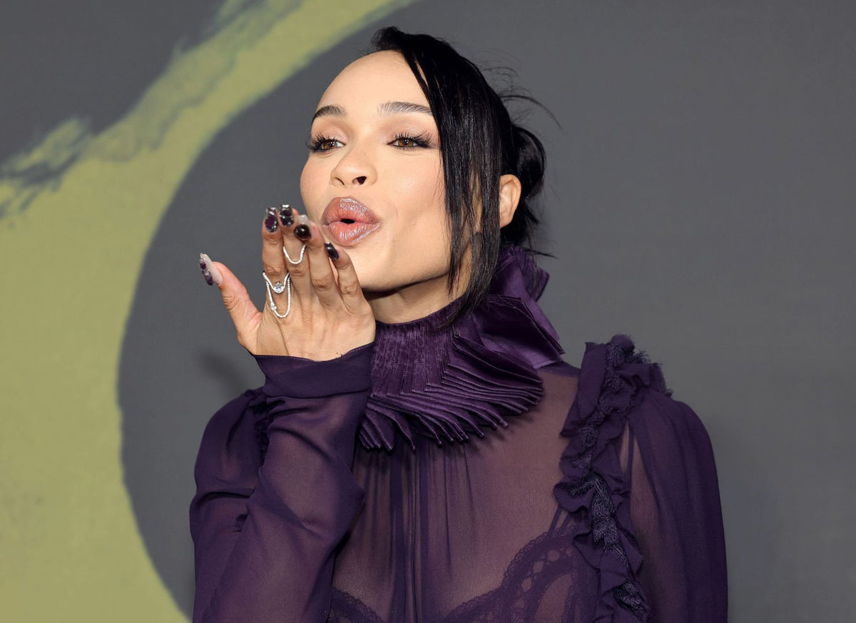Cleopatra Coleman blows a kiss while wearing a sculptural silver ring and multiple statement rings. She pairs her jewelry with a sheer purple blouse featuring a ruffled high collar and dramatic sleeves.