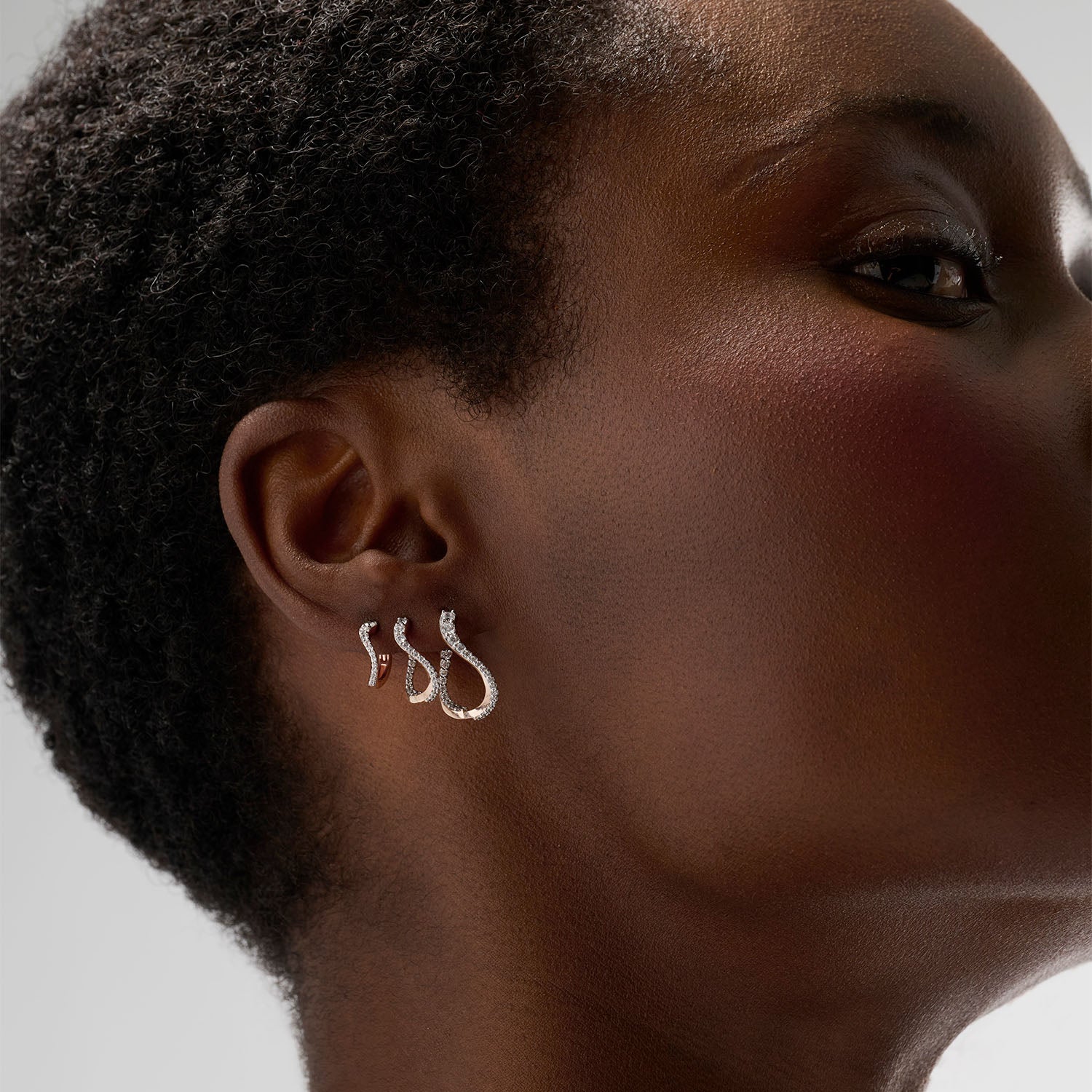 A young woman wearing three curved hoop earrings in a gradient sizes. Each hoop is set with diamonds and crafted in 18k rose gold. Timeless and luminous, this set and its undulating curves evoke caustic light, blending MARIE MAS’s two signature sources of inspiration: water and light.