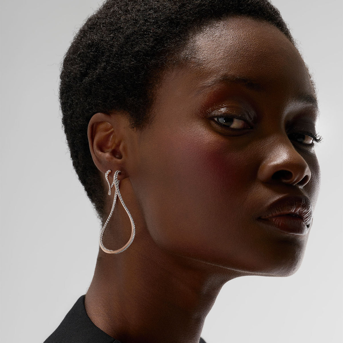 A young woman is wearing two hoop earrings, one of which is a transformable bangle-hoop earring. The fine jewelry pieces are crafted in 18k gold and set with natural diamonds.