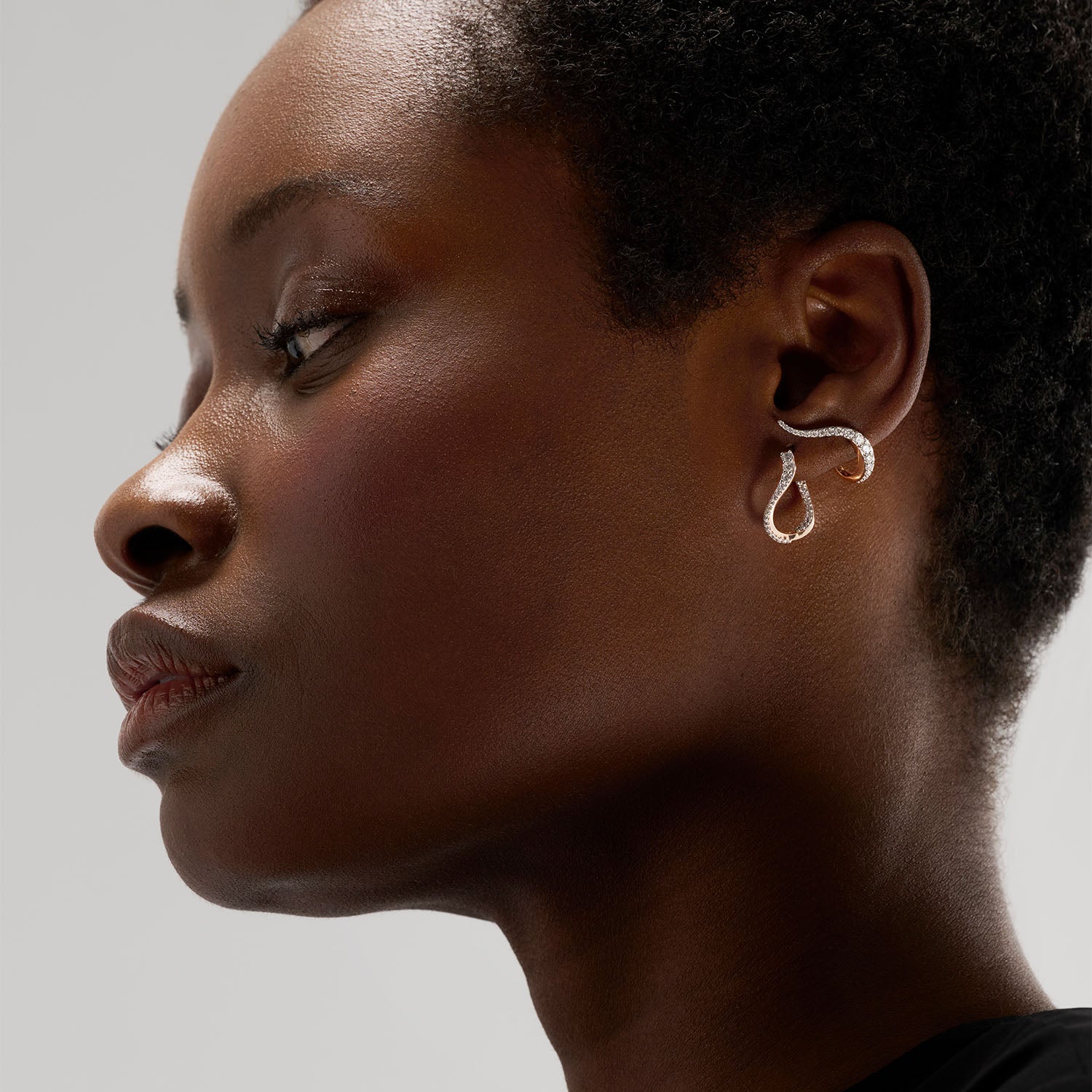 A young woman wear an ear set composed of a hoop earring and an earring. The pieces draw luminous lines on the ears, reminding the caustic light movement.