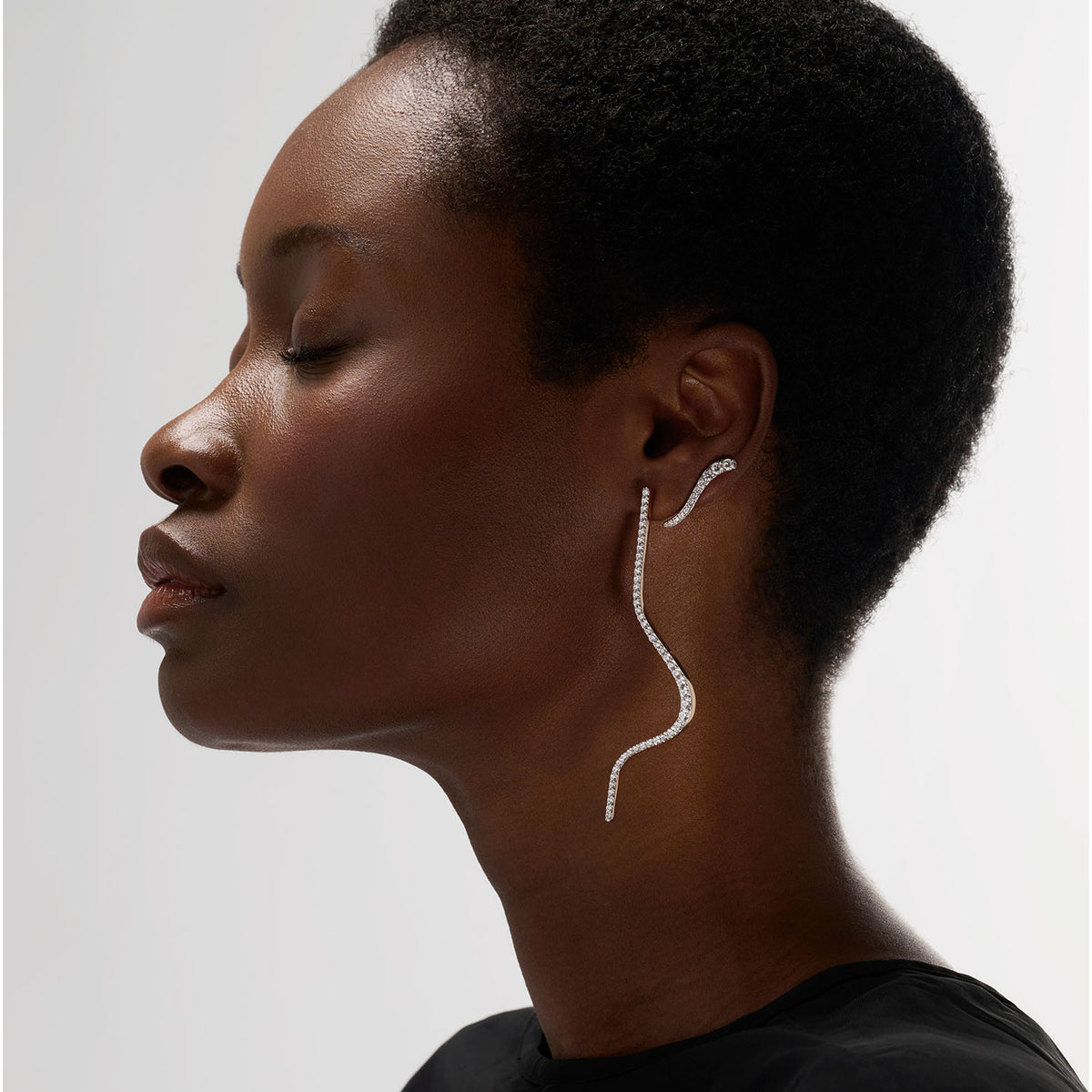 A woman wearing a transformable jewel brooch-earring featuring an ingenious mechanism that allows the posts to retract or extend, ensuring a secure and comfortable wear whether styled on the ear or as a brooch. This fine jewelry piece is crafted in 18k gold and set with natural diamonds. Its curves evoke caustic light, where water and light meet.