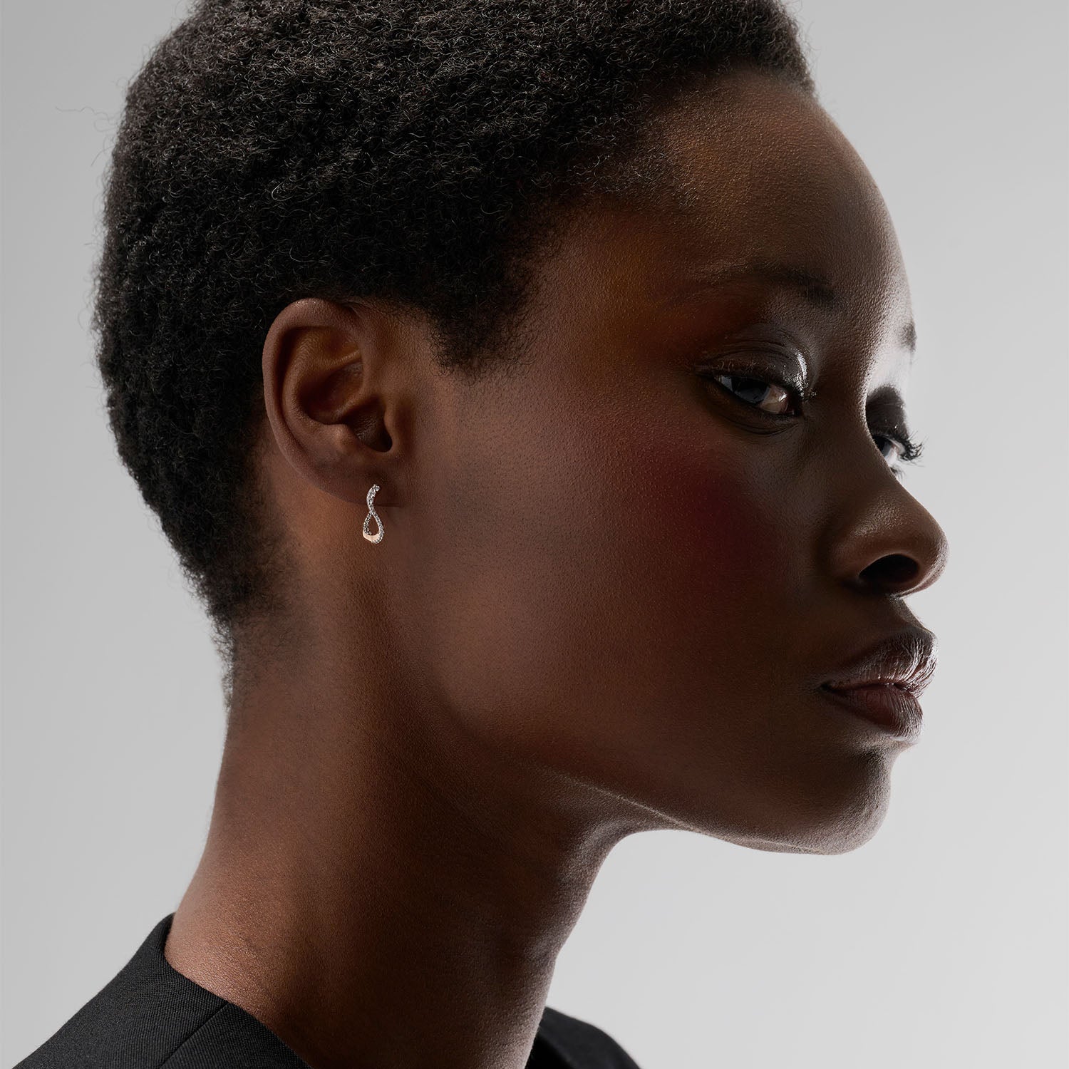 A young woman wearing a petite hoop earring. This piece is crafted in 18k rose gold and set with natural diamonds. This jewel and its undulations are inspired by the Caustics, the luminous phemonem of light meeting water.