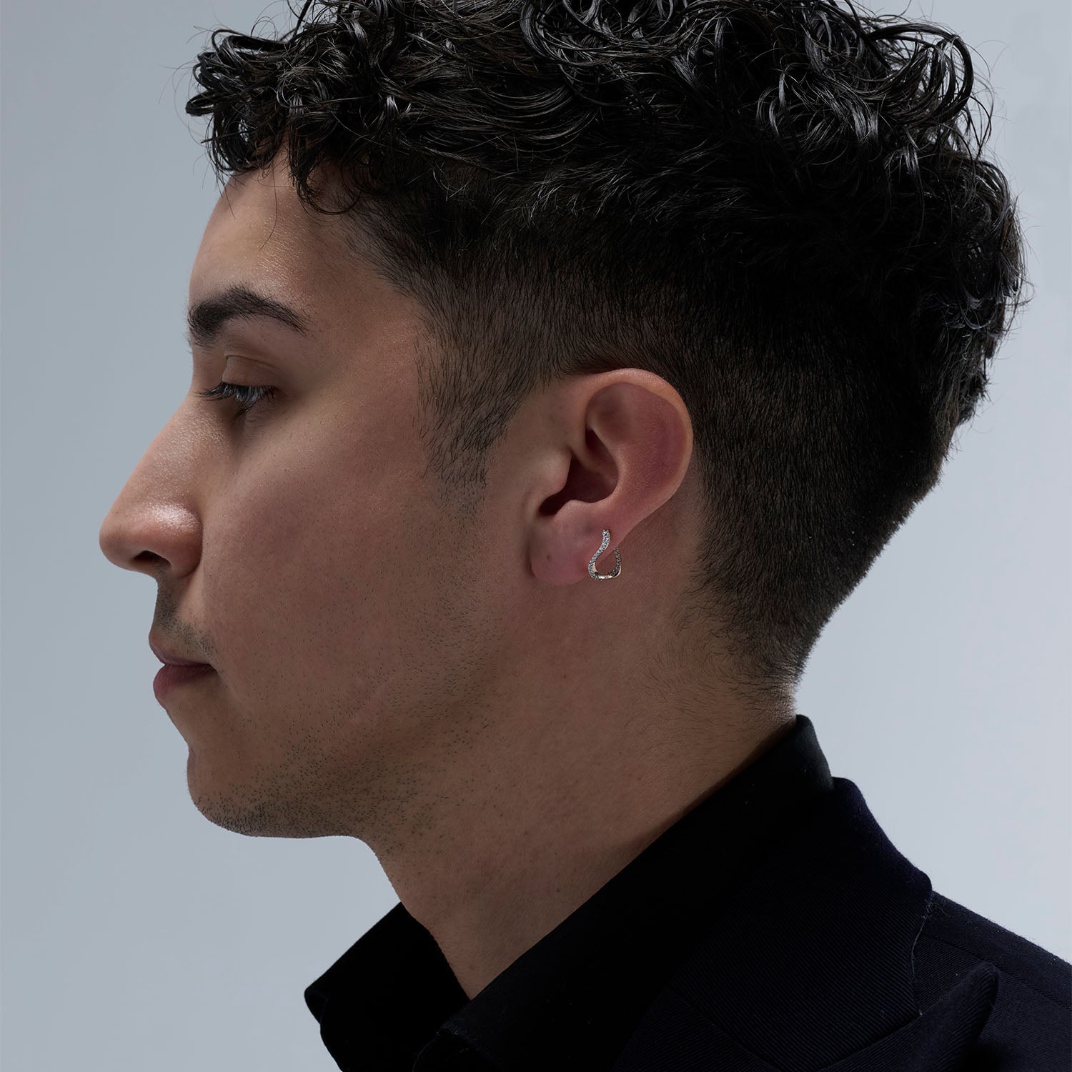 A young man wearing a petite hoop earring as piercing. This piece is crafted in 18k rose gold and set with natural diamonds. This jewel and its undulations are inspired by the Caustics, the luminous phemonem of light meeting water.