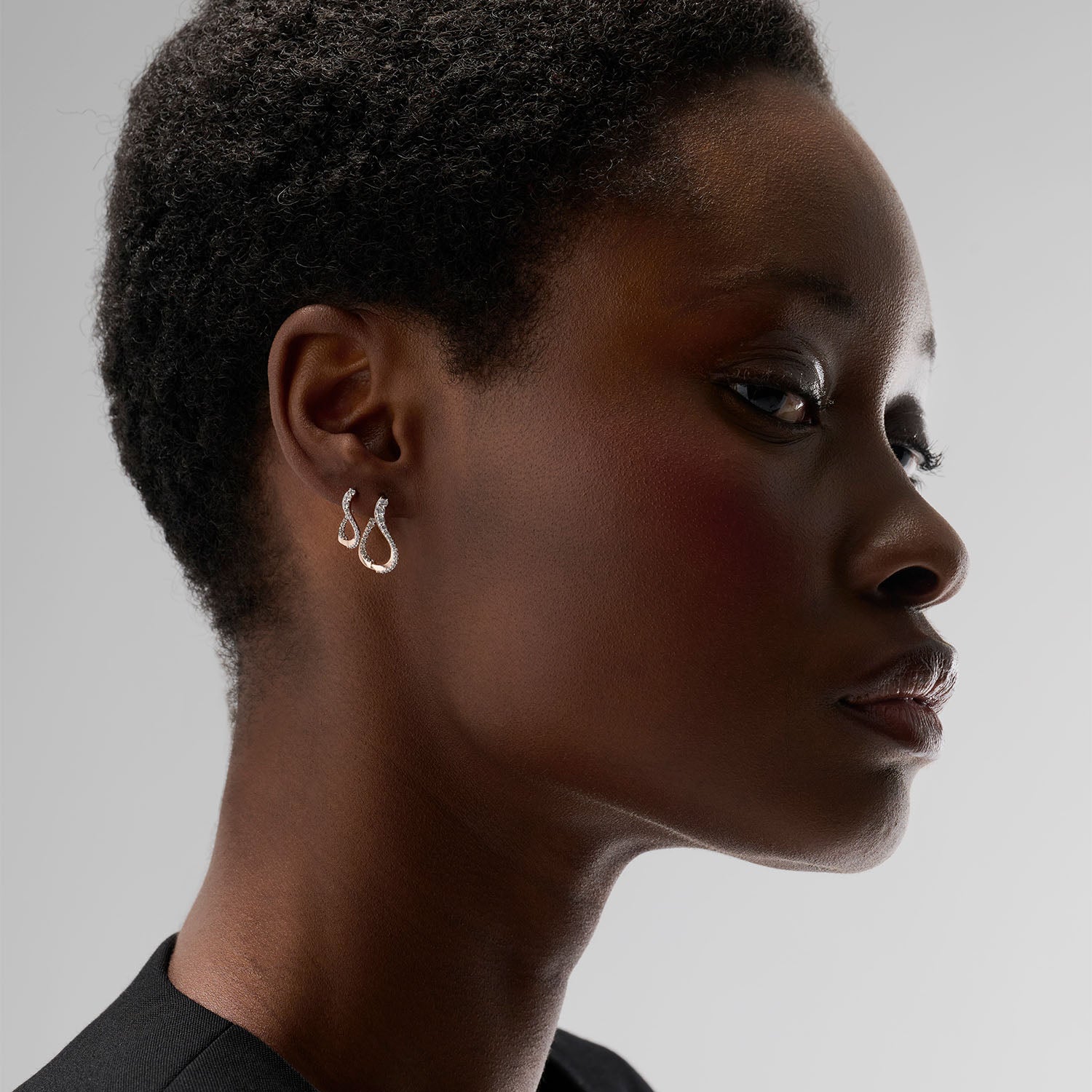 A young woman wearing a set composed of two curved hoop earrings. Each pieces is crafted in 18k rose gold and set with natural diamonds. Evoking the caustic light, they draw luminous lines around the earlobe that ondulate thanks to their curves.