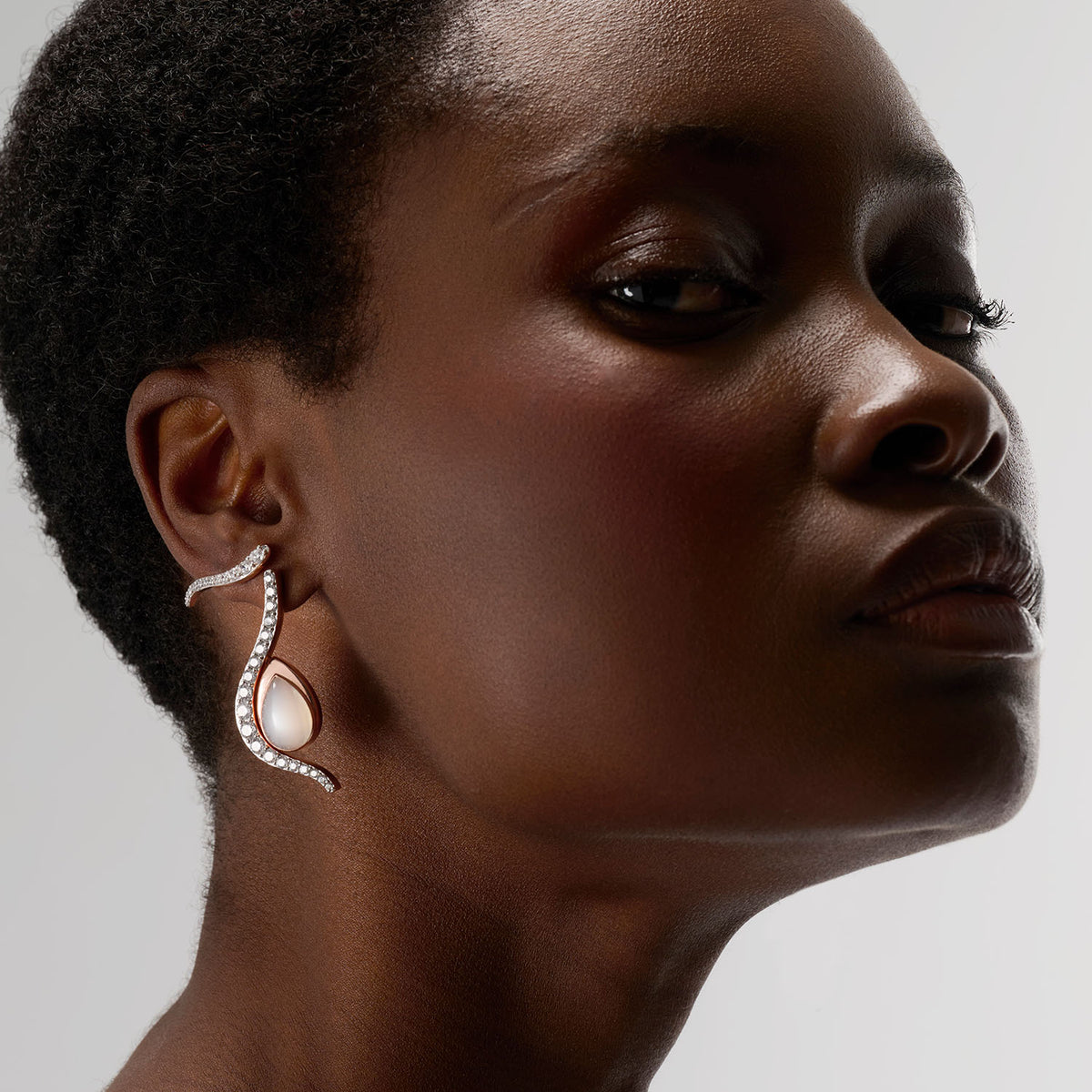 A young woman wearing an ear set composed of thrre brooch-earrings. Crafted in 18k gold and set with natural diamonds or a white moonstone, the curves remind the water and the undulation of the Causics. Thanks to an ingenious system, the white moonstone piece can be attached to or detached from the earring to create a multitude of looks.