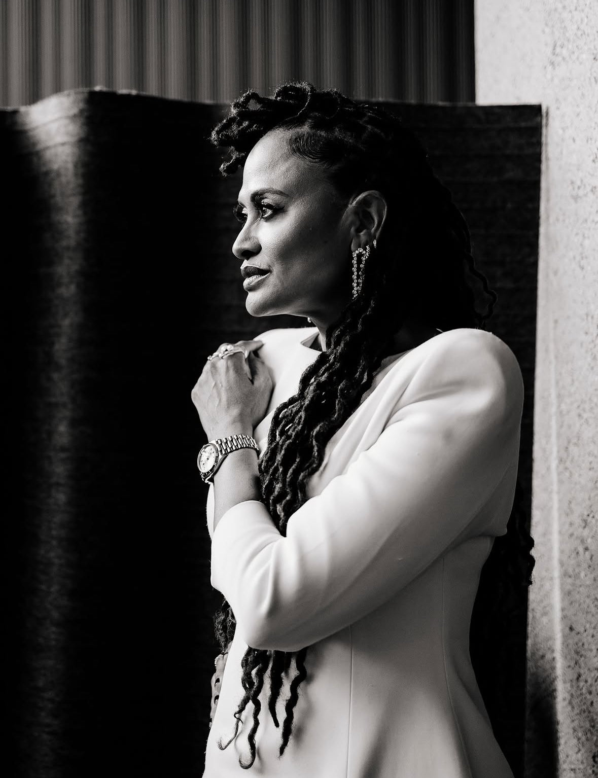 Ava DuVernay poses in profile at the New York Film Festival, wearing a white tailored dress with long braids draped over one shoulder, and rings. The black-and-white portrait highlights her jewelry against a contrasting architectural backdrop.