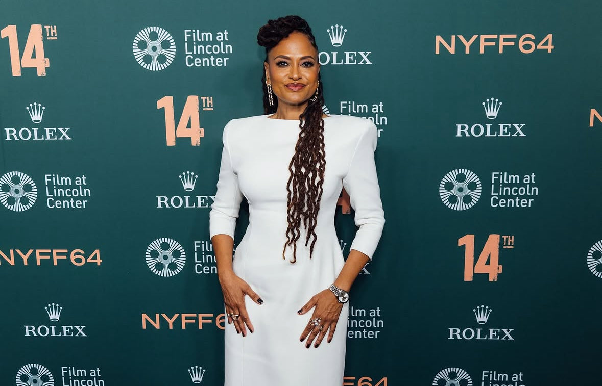 Ava DuVernay attends the 64th New York Film Festival in a fitted white dress with structured shoulders, and long braids. She accessorizes with multiple statement rings, posing against the teal Film at Lincoln Center and Rolex press backdrop.