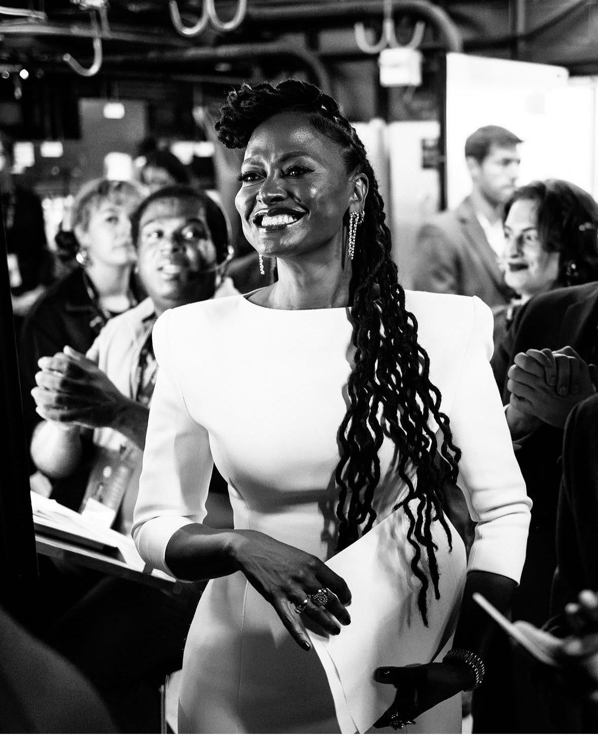 Ava DuVernay smiles during the New York Film Festival, wearing a structured white dress with three-quarter sleeves, long braided hair, and statement rings. Photographers and attendees surround her as she interacts with the crowd in this black-and-white event photograph.