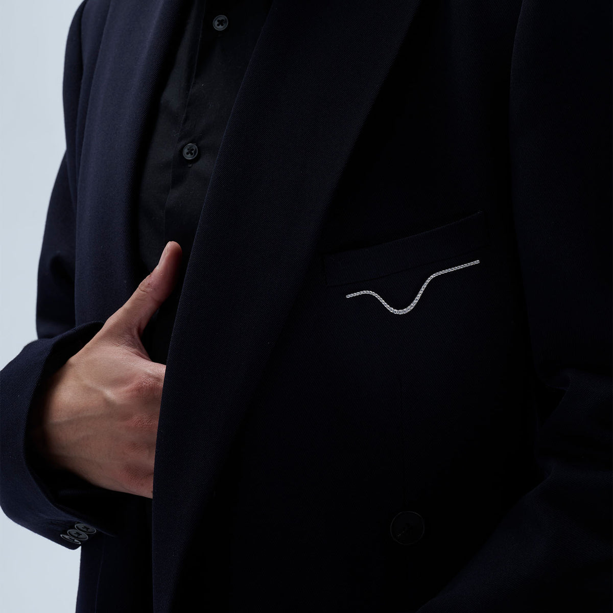 A man wearing a transformable jewel brooch-earring featuring an ingenious mechanism that allows the posts to retract or extend, ensuring a secure and comfortable wear whether styled on the ear or as a brooch. This fine jewelry piece is crafted in 18k gold and set with natural diamonds. Its curves evoke caustic light, where water and light meet.