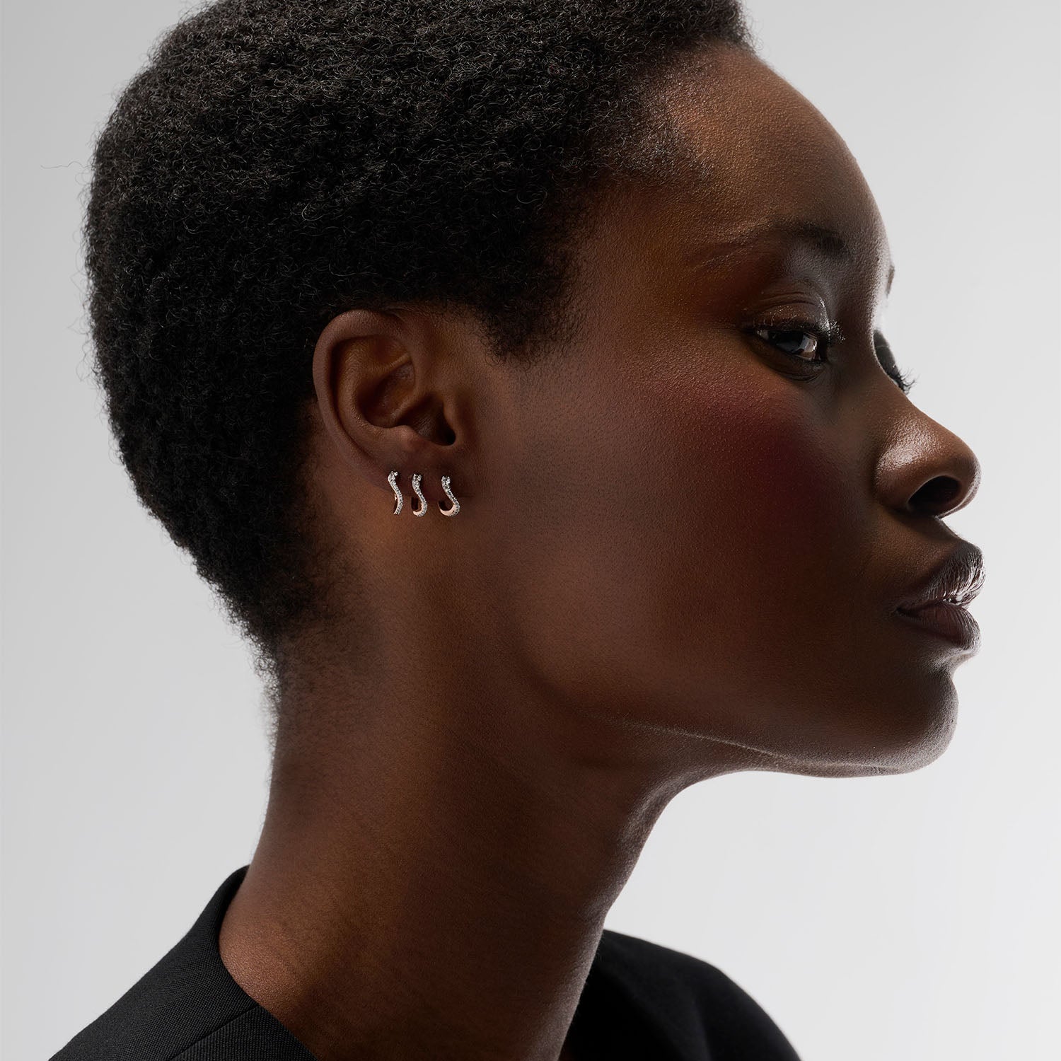 A young woman wearing a set of three identical pieces: mini hoop earrings set with natural diamonds and crafted in 18k rose gold. These understated hoops bring a touch of originality through their delicate curve, subtly evoking the refined movement of caustic light.