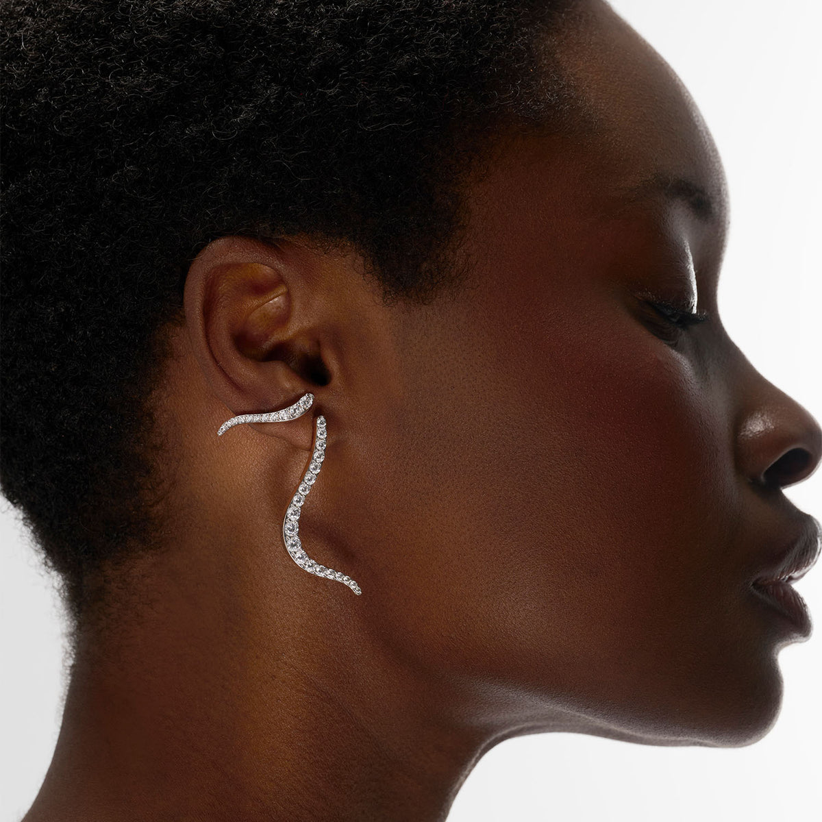 A woman wearing a transformable jewel brooch-earring as earring featuring an ingenious mechanism that allows the posts to retract or extend, ensuring a secure and comfortable wear whether styled on the ear or as a brooch. This fine jewelry piece is crafted in 18k gold and set with natural diamonds. Its curves evoke caustic light, where water and light meet.