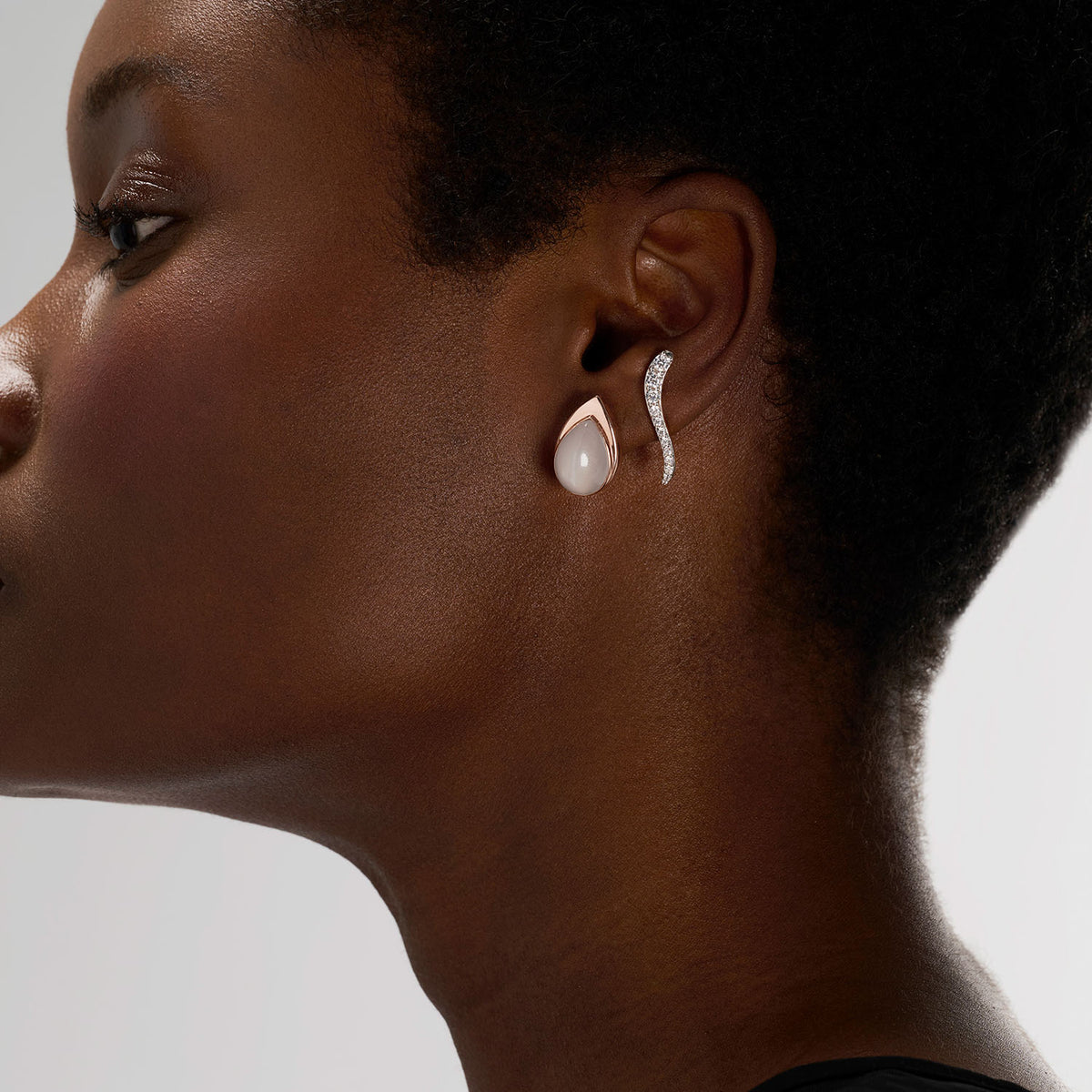 A young woman wearing a set of two pieces composed of an earring, set with natural diamonds, and a transformable jewel brooch-earring, set with a pear white moonstone. The fine jewelry pieces are crafted in 18k rose gold and inspired by the meeting of light and water.