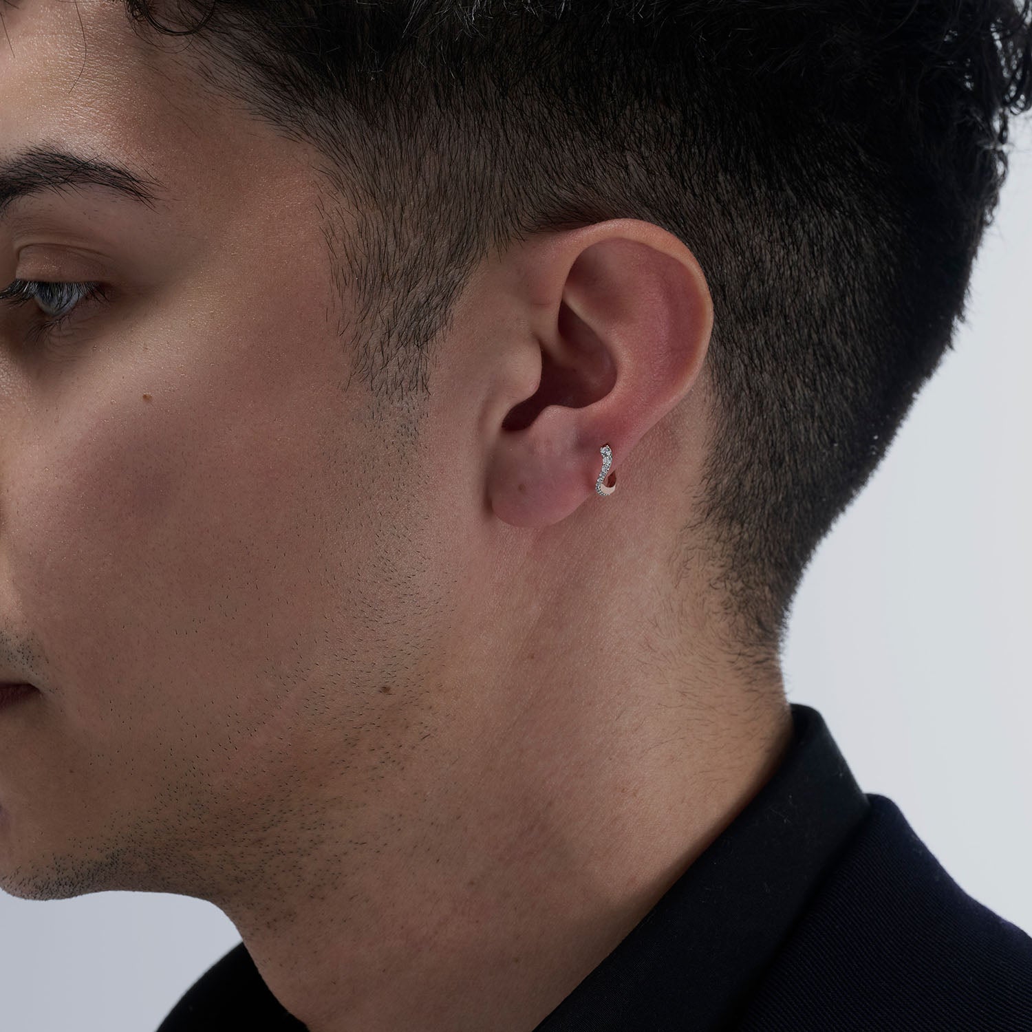 A young man wearing a mini hoop earring as piercing. This piece is crafted in 18k rose gold and set with natural diamonds. This jewel and its undulations are inspired by the Caustics, the luminous phemonem of light meeting water.