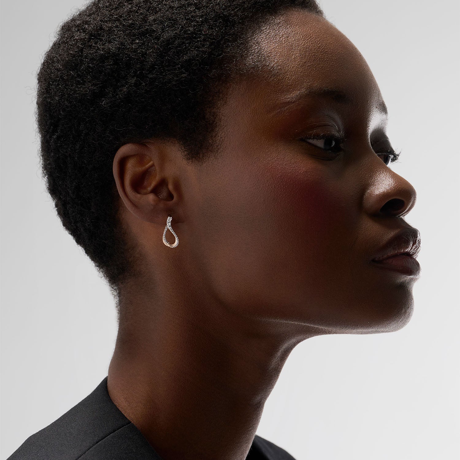 A young woman wearing a beautiful undulating hoop earring. This piece is crafted in 18k rose gold and set with natural diamonds. This jewel and its undulations are inspired by the Caustics, the luminous phemonem of light meeting water.