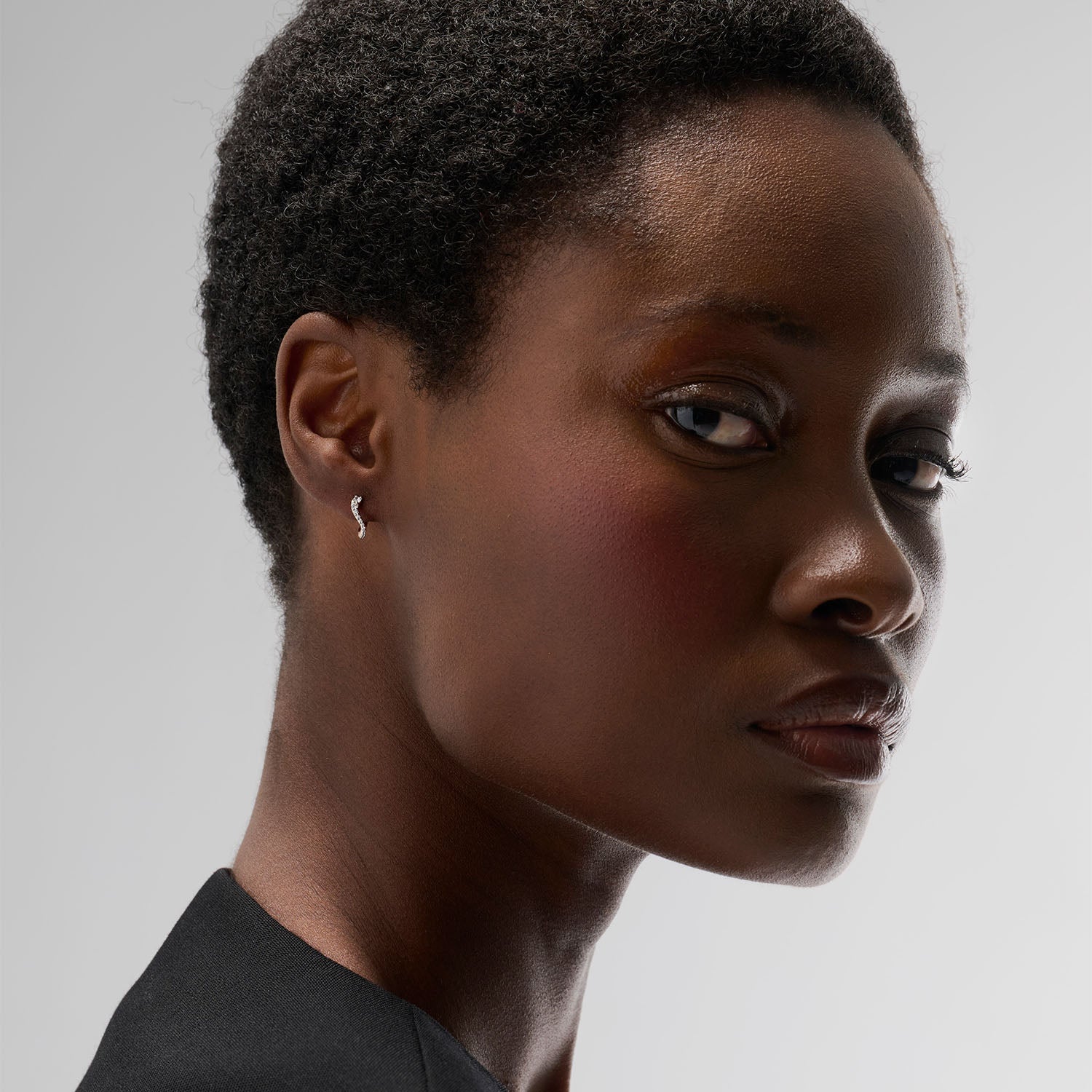 A young woman wearing a mini hoop earring. This piece is crafted in 18k rose gold and set with natural diamonds. This jewel and its undulations are inspired by the Caustics, the luminous phemonem of light meeting water.