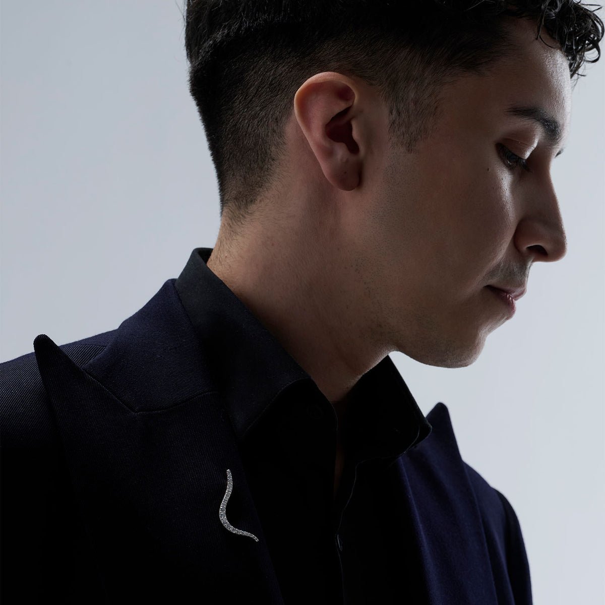 A man wearing a transformable jewel brooch-earring featuring an ingenious mechanism that allows the posts to retract or extend, ensuring a secure and comfortable wear whether styled on the ear or as a brooch. This fine jewelry piece is crafted in 18k gold and set with natural diamonds. Its curves evoke caustic light, where water and light meet.