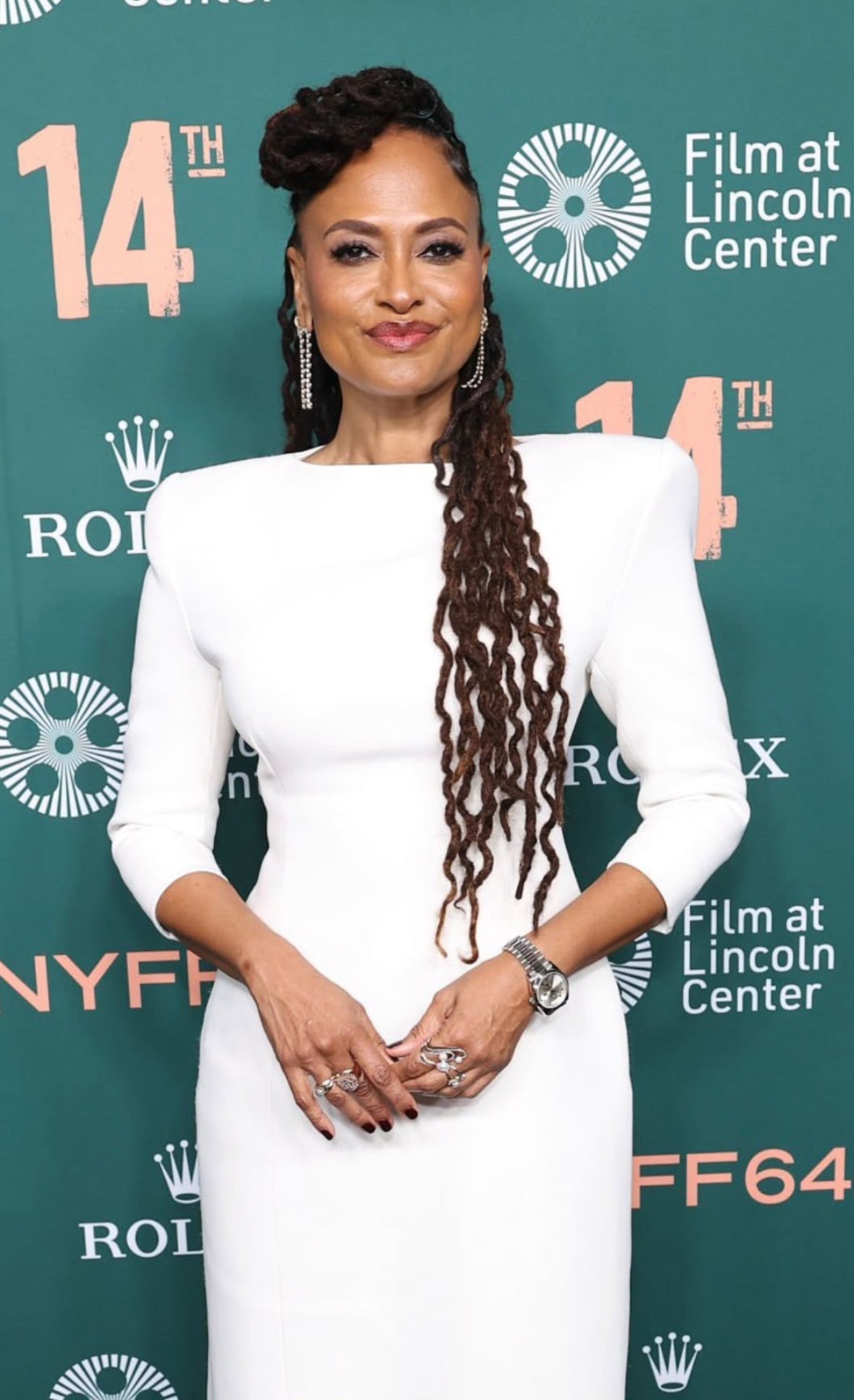 Ava DuVernay attends the New York Film Festival in a fitted white dress with structured shoulders, accessorized with rings. Her long braids fall over one shoulder as she poses against the teal NYFF and Film at Lincoln Center event backdrop.